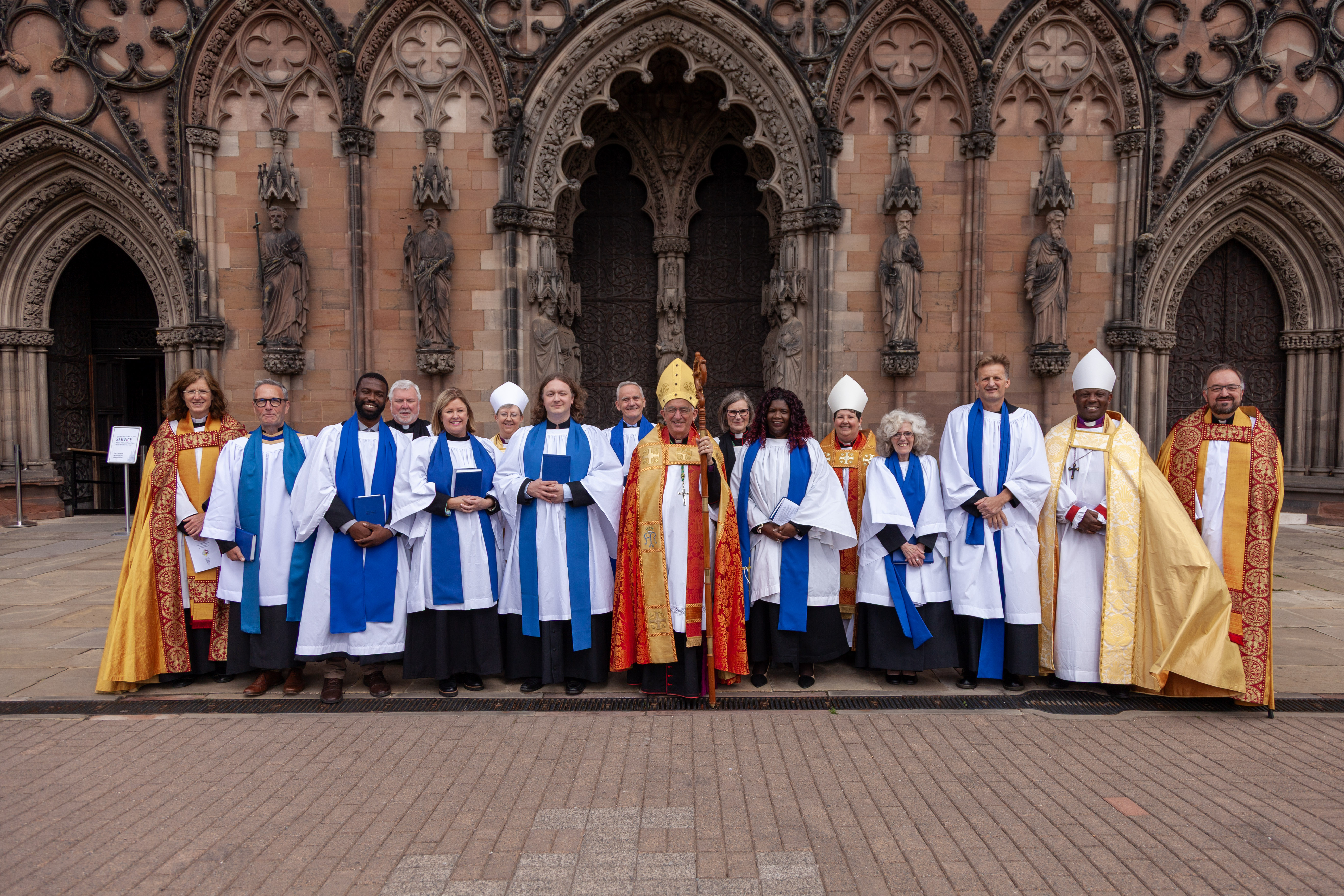Seven newly-licensed Readers standing outside the west door of Lichfield Cathedral, with Bishop Michael Ipgrave in the middle and surrounded by accompanying bishops and archdeacons